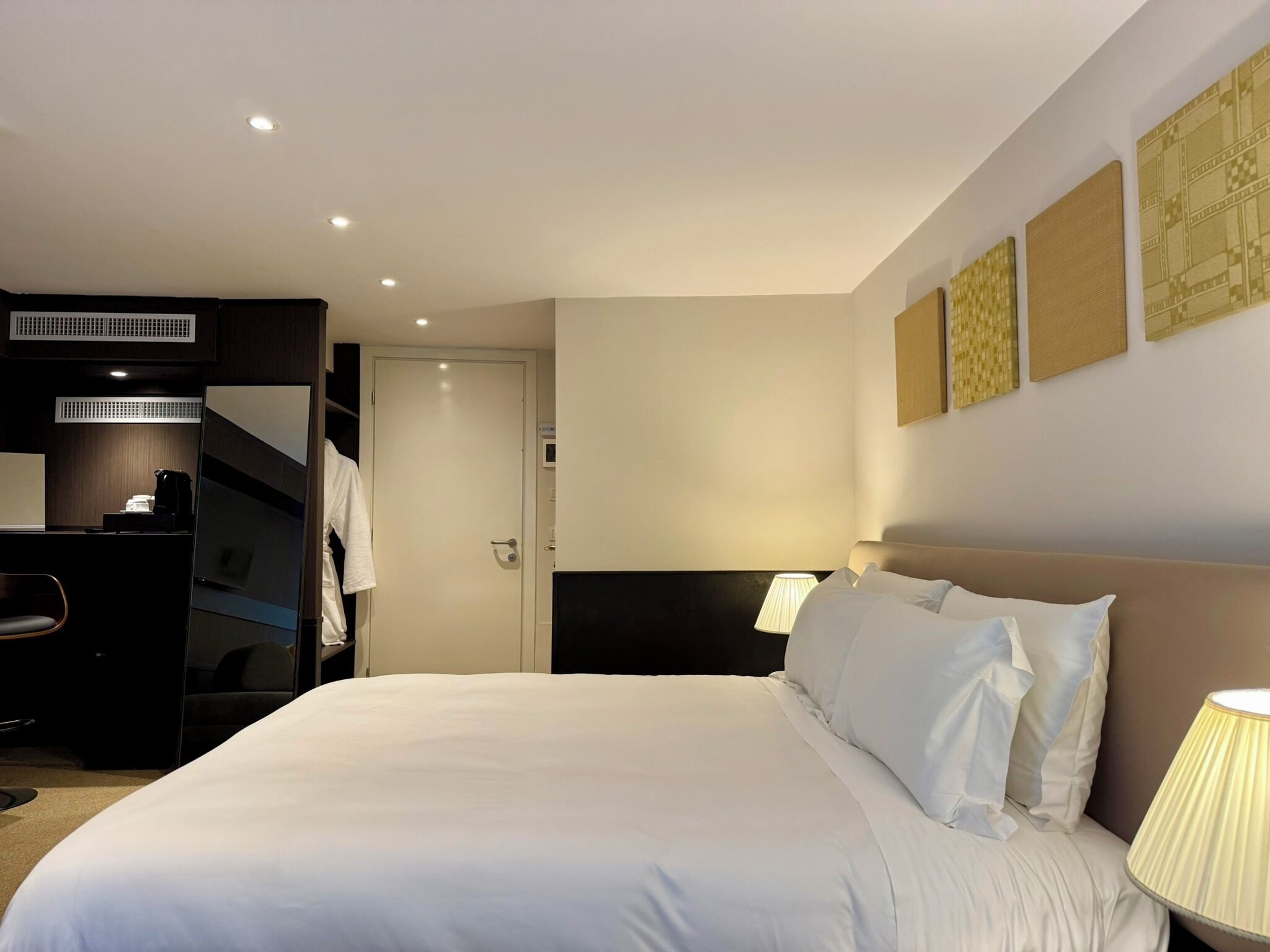 Quarto de hotel com cama, luminárias de cabeceira, uma escrivaninha e um guarda-roupa.