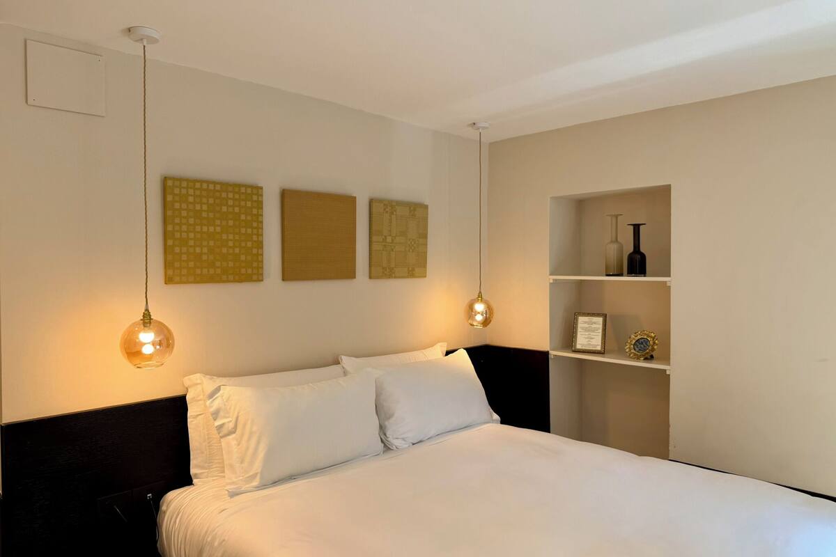 A neatly made bed with white linens, a dark headboard, and two pendant lights. There are three framed artworks on the wall above the bed.
