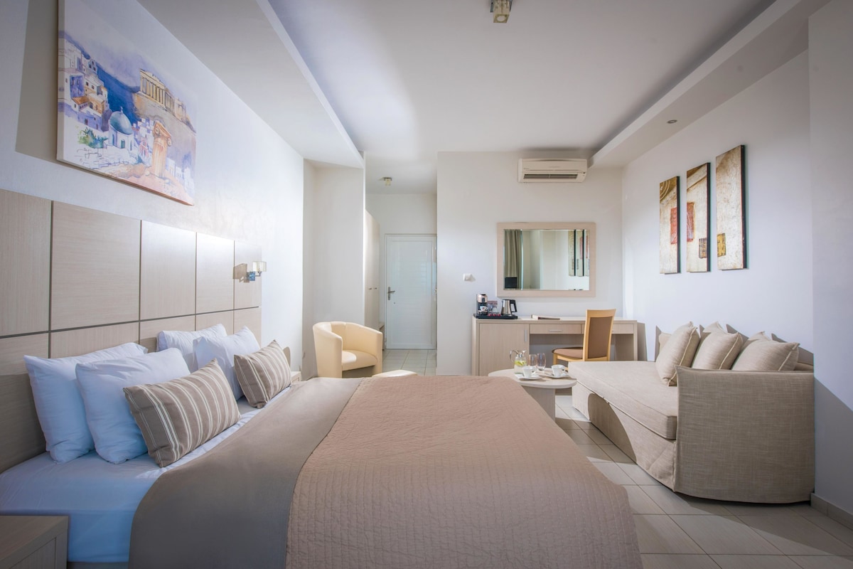 Ein modernes Hotelzimmer mit einem großen Bett, einer Sitzecke, einem Schreibtisch und einem Sessel. An der Wand hängt ein Gemälde und über dem Schreibtisch befindet sich ein Spiegel.