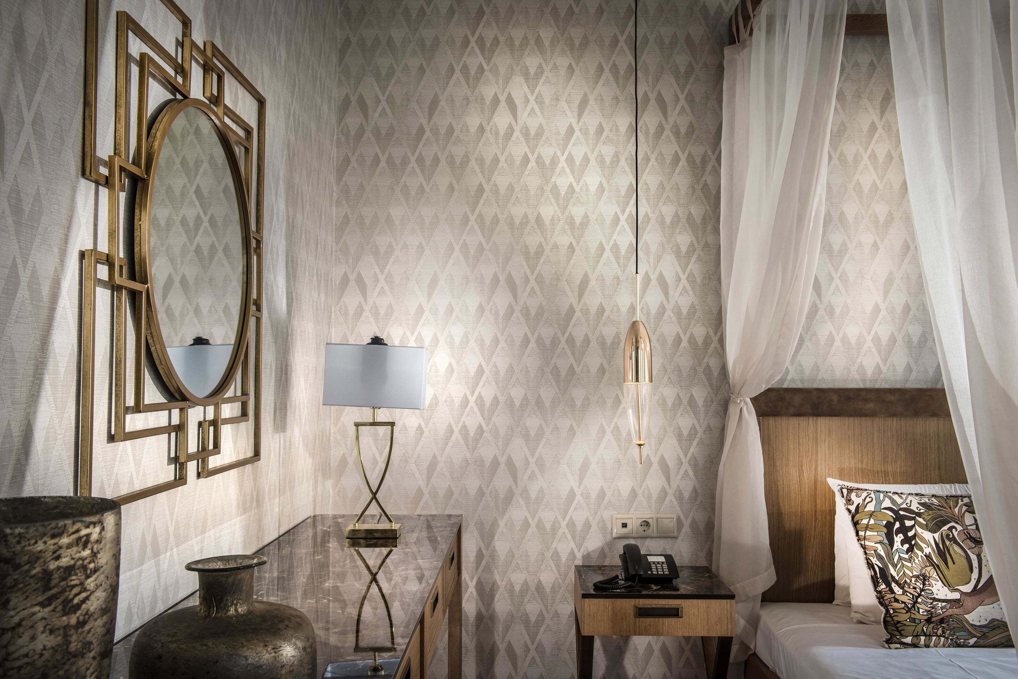 Una habitación de hotel con cama, mesita de noche, lámpara, teléfono y un espejo decorativo.