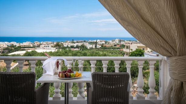 Een balkon met rieten stoelen, een tafel met fruit en een fles, en uitzicht op zee en de stad.