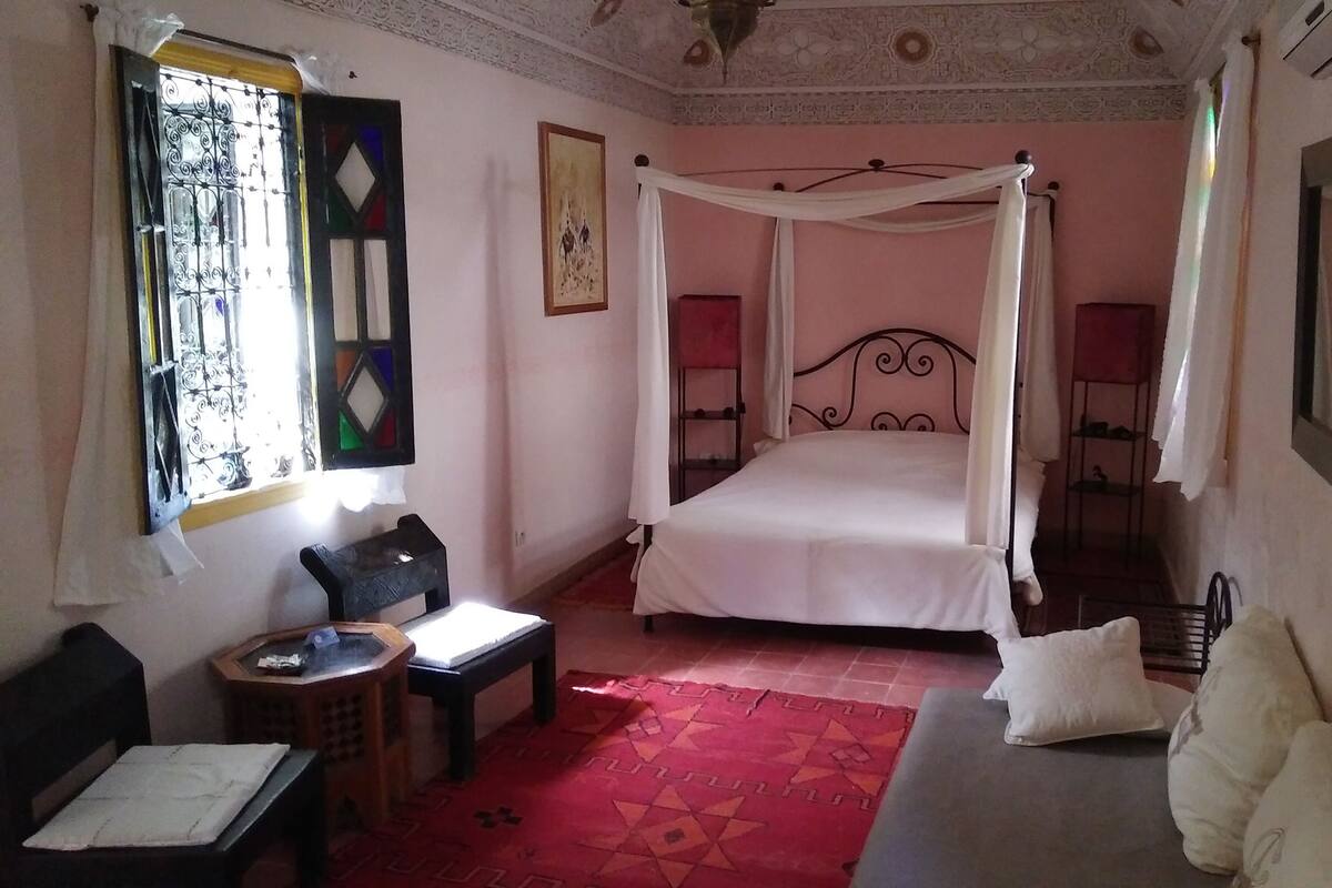 A bedroom with a four-poster bed, a small table, a chair, and a bedside lamp.