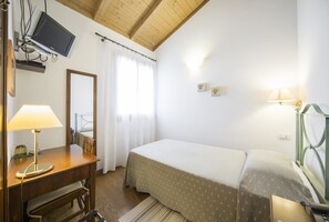 Un dormitorio con cama, escritorio con lámpara, televisión montada en la pared y ventana con cortinas.