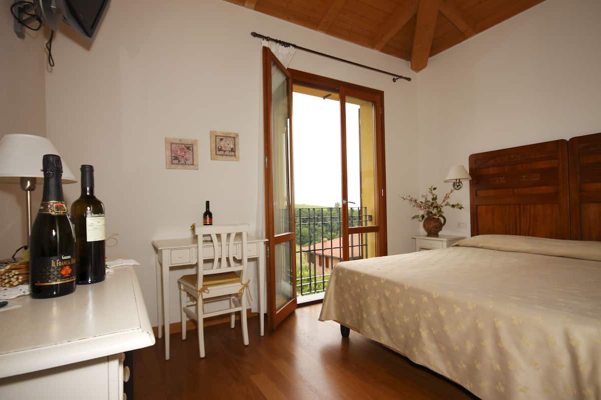 A bedroom with a bed, a desk with two bottles, a chair, a small table with a vase, and a balcony with a railing.