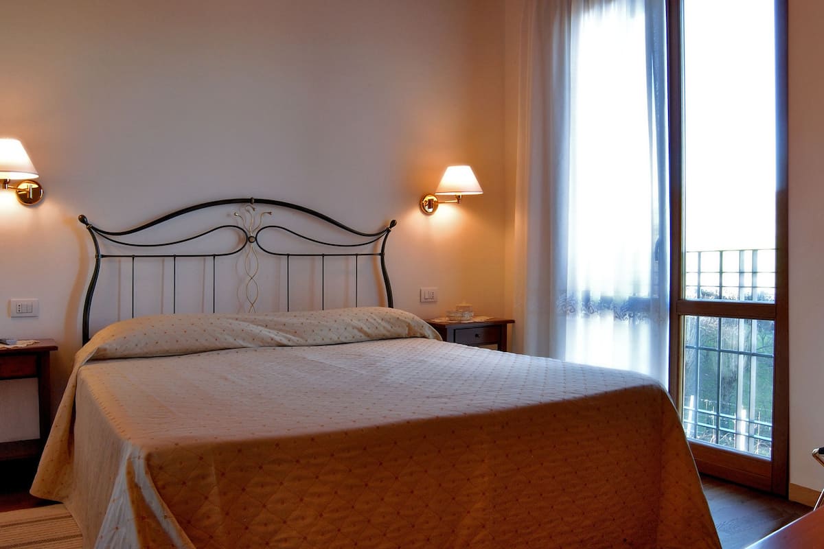 A hotel room with a bed, bedside lamps, a table with a wine bottle and glasses, and a view through a window.