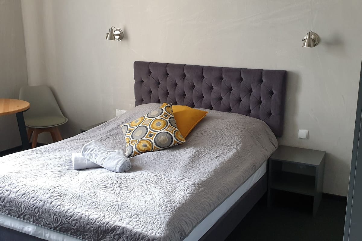 Uma cama bem arrumada com cabeceira cinza, almofada estampada e uma mesinha de cabeceira.