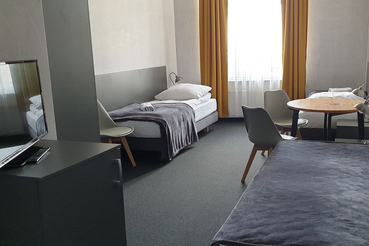 Quarto de hotel com cama, escrivaninha com cadeira, mesa pequena com duas cadeiras, televisão e janela com cortinas.