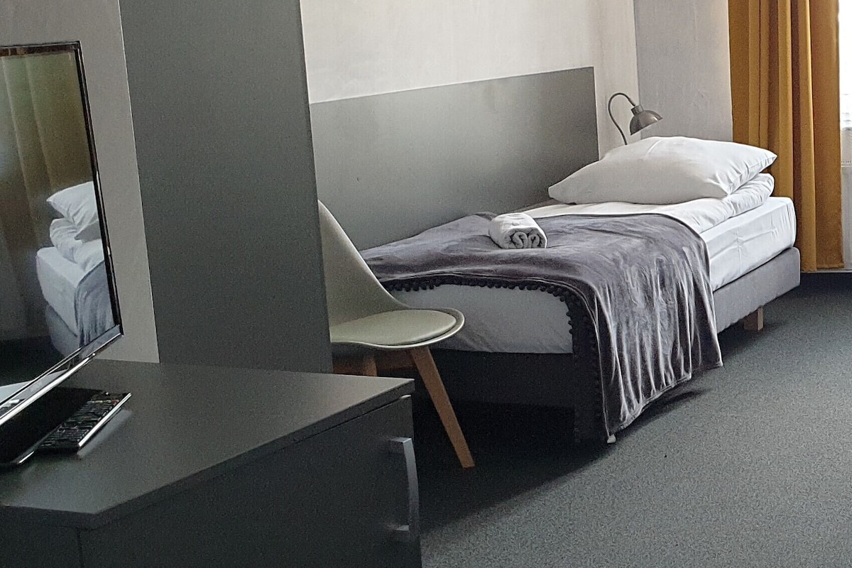 Quarto de hotel com cama, escrivaninha com computador e uma cadeira.