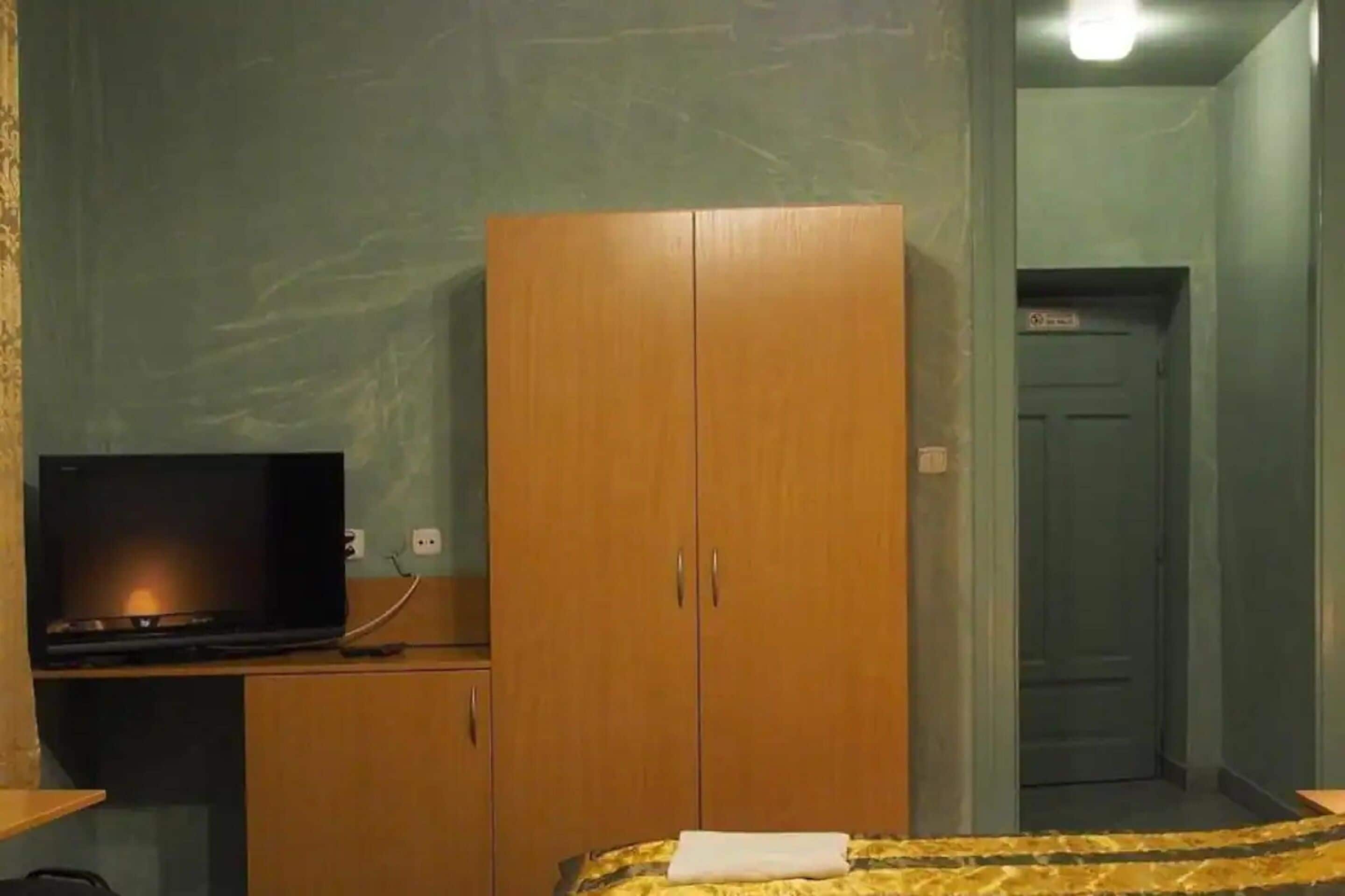Une pièce comprenant un lit, une armoire en bois, un téléviseur posé sur un bureau et une porte donnant sur une autre pièce.
