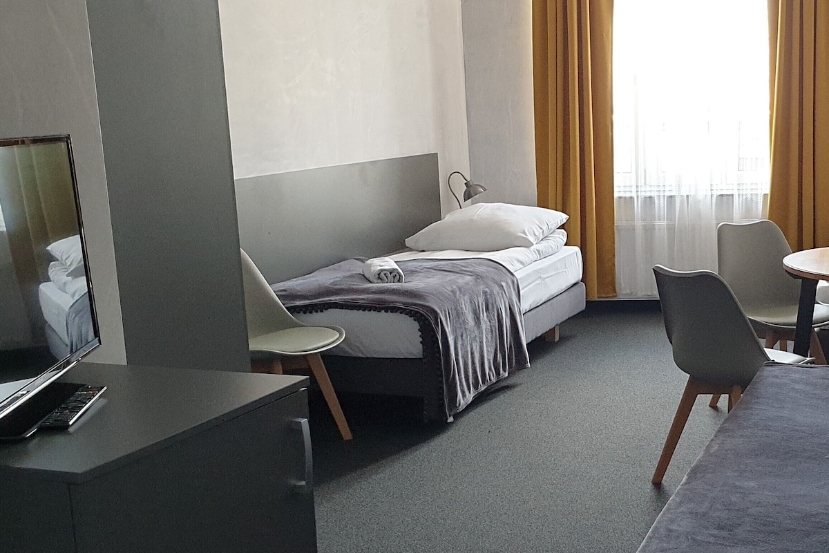 Quarto de hotel com cama, escrivaninha, um laptop, cadeira e uma pequena mesa.