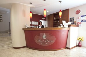 A reception desk at the San Marino Hotel with a receptionist, a clock, and a vase with sunflowers. - Hotel San Martino (Syracuse)