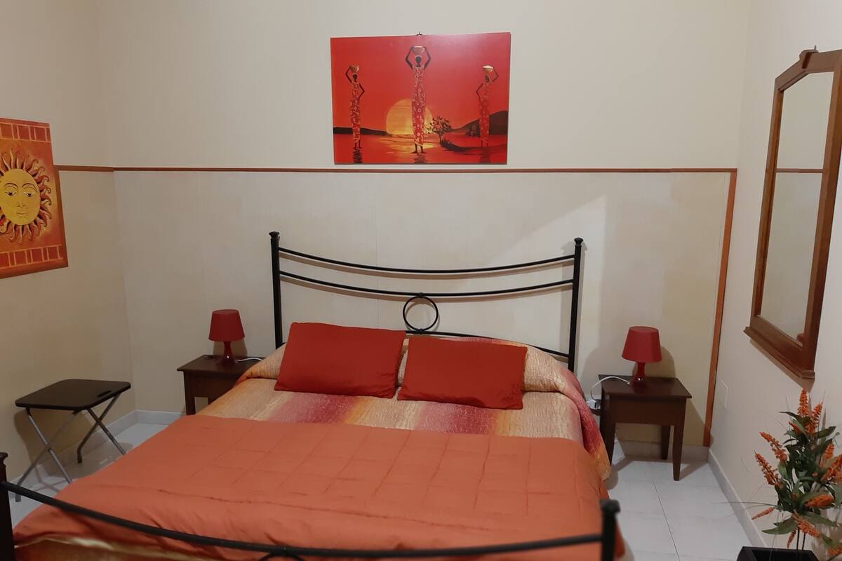 A neatly arranged bedroom with a metal bed frame, two red lamps, a red and orange bedspread, a wall-mounted mirror, and a framed painting above the bed.
