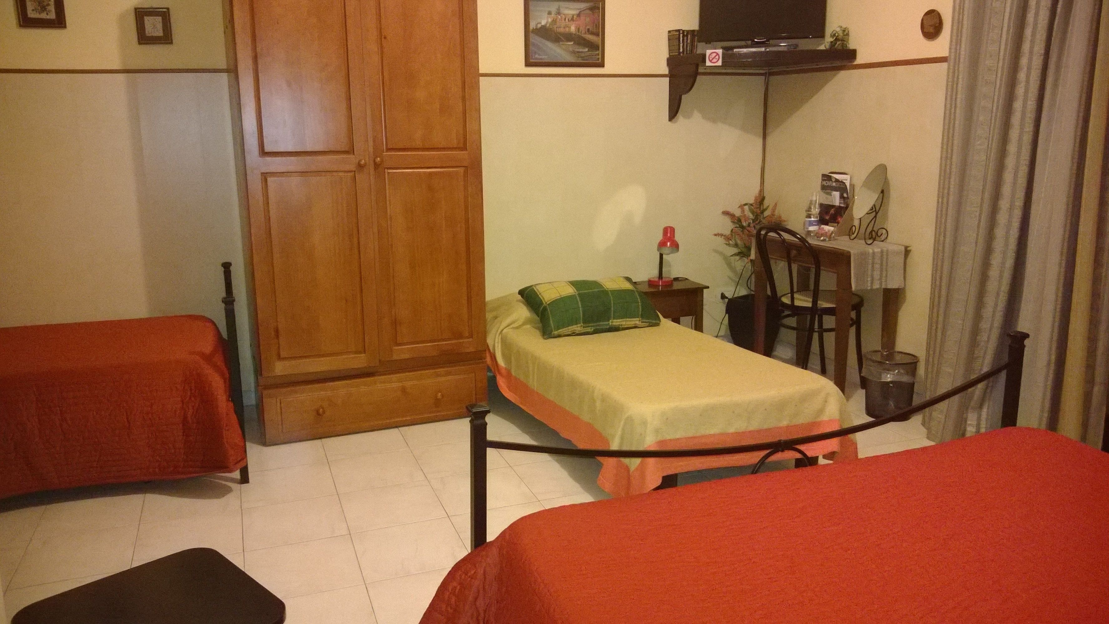 A room with two single beds, a wooden wardrobe, a desk with a chair, a television, and a small table with a plant.