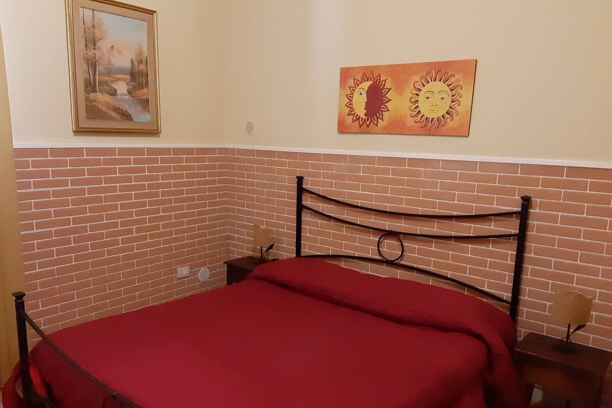 A room with a brick-patterned wall, a bed with a red bedspread, a nightstand, a lamp, and a painting on the wall.