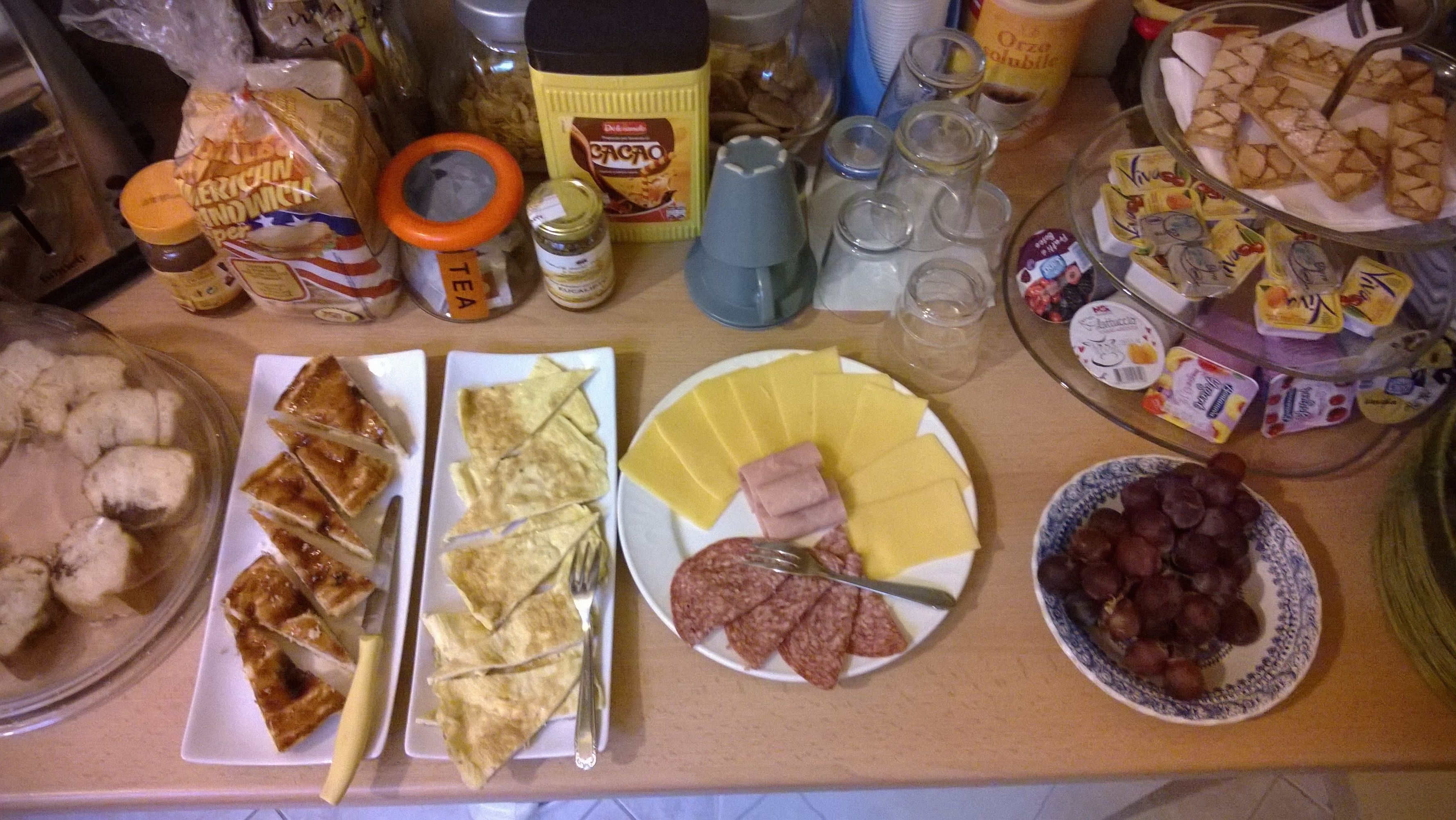 A table with various food items and condiments, including plates of sliced bread, cheese, and meats, as well as jars of preserves and condiments.