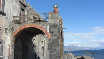 Une arche en pierre, surmontée d’une rambarde métallique et d’un pot de fleurs, mène à une zone côtière où le ciel est dégagé et où l’on aperçoit des montagnes à l’horizon.