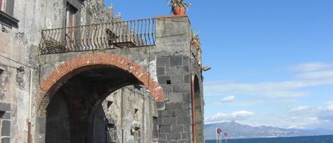 An arched stone gateway with a metal railing and a potted plant on top, leading to a coastal area with a clear sky and mountains in the background.