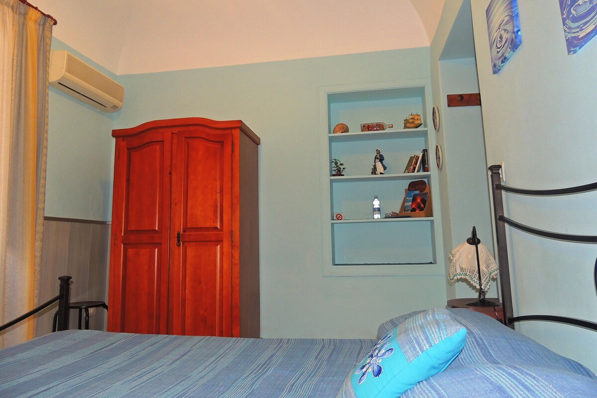 A bedroom with a bed, a wardrobe, a shelf with decorative items, and a window with curtains.