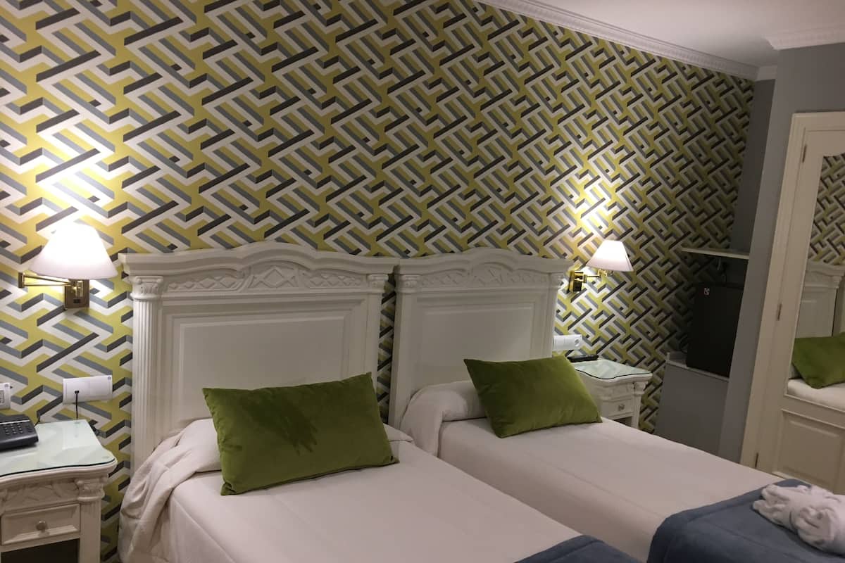 Une chambre d’hôtel avec deux lits, chacun agrémenté d’un oreiller vert, d’une tête de lit blanche et de tables de chevet avec des lampes. Les murs sont recouverts d’un papier peint à motifs géométriques.