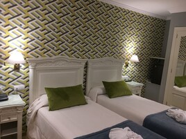 Habitación de hotel con dos camas, cada una con almohada verde, cabeceros blancos y mesitas de noche con lámparas. Las paredes tienen un papel pintado con estampado geométrico.