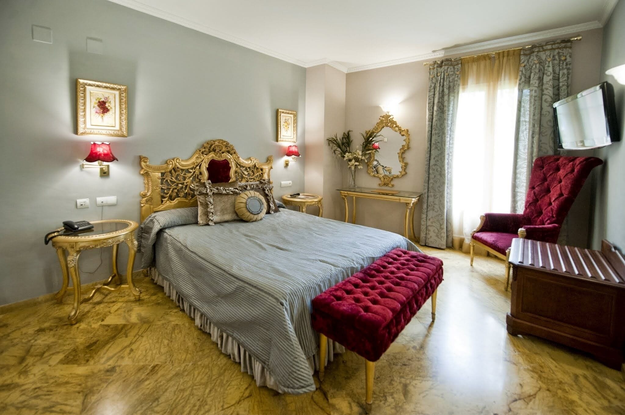 A bedroom with a large bed, a red armchair, a wooden nightstand, a gold-framed mirror, and a television mounted on the wall.