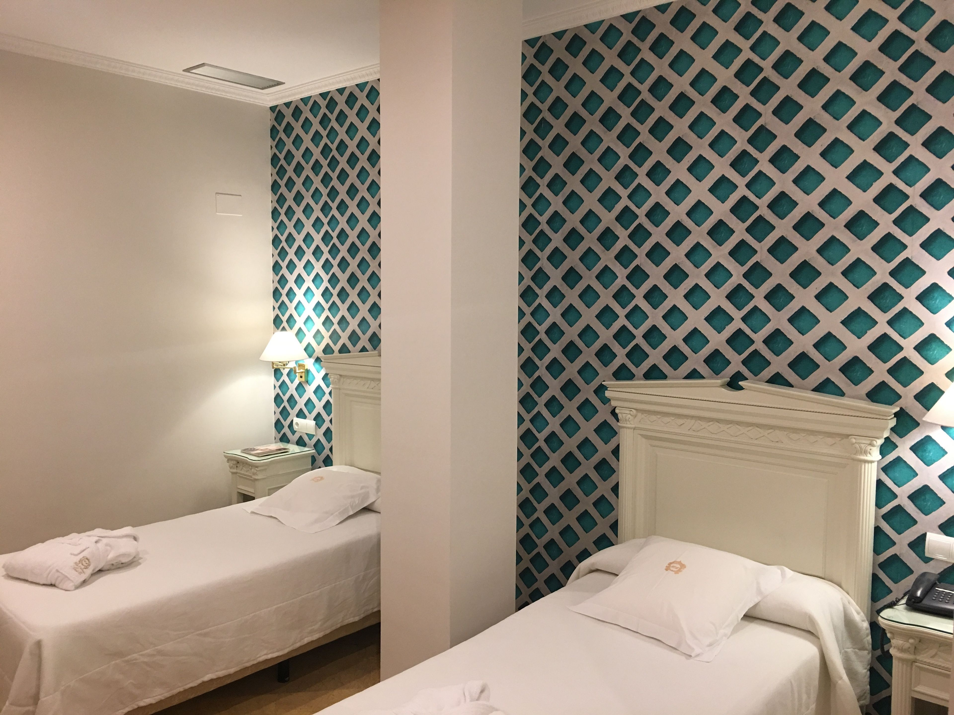 A hotel room with two single beds, a nightstand with a lamp, and a patterned wall.