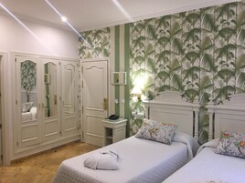 Habitación de hotel con dos camas, un armario, una mesita de noche con lámpara y una pared con diseño de hojas tropicales.