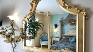 Una habitación con un espejo grande y ornamentado, una mesa con cubierta de vidrio y un cuenco decorativo, un florero con flores y un cuadro en la pared.