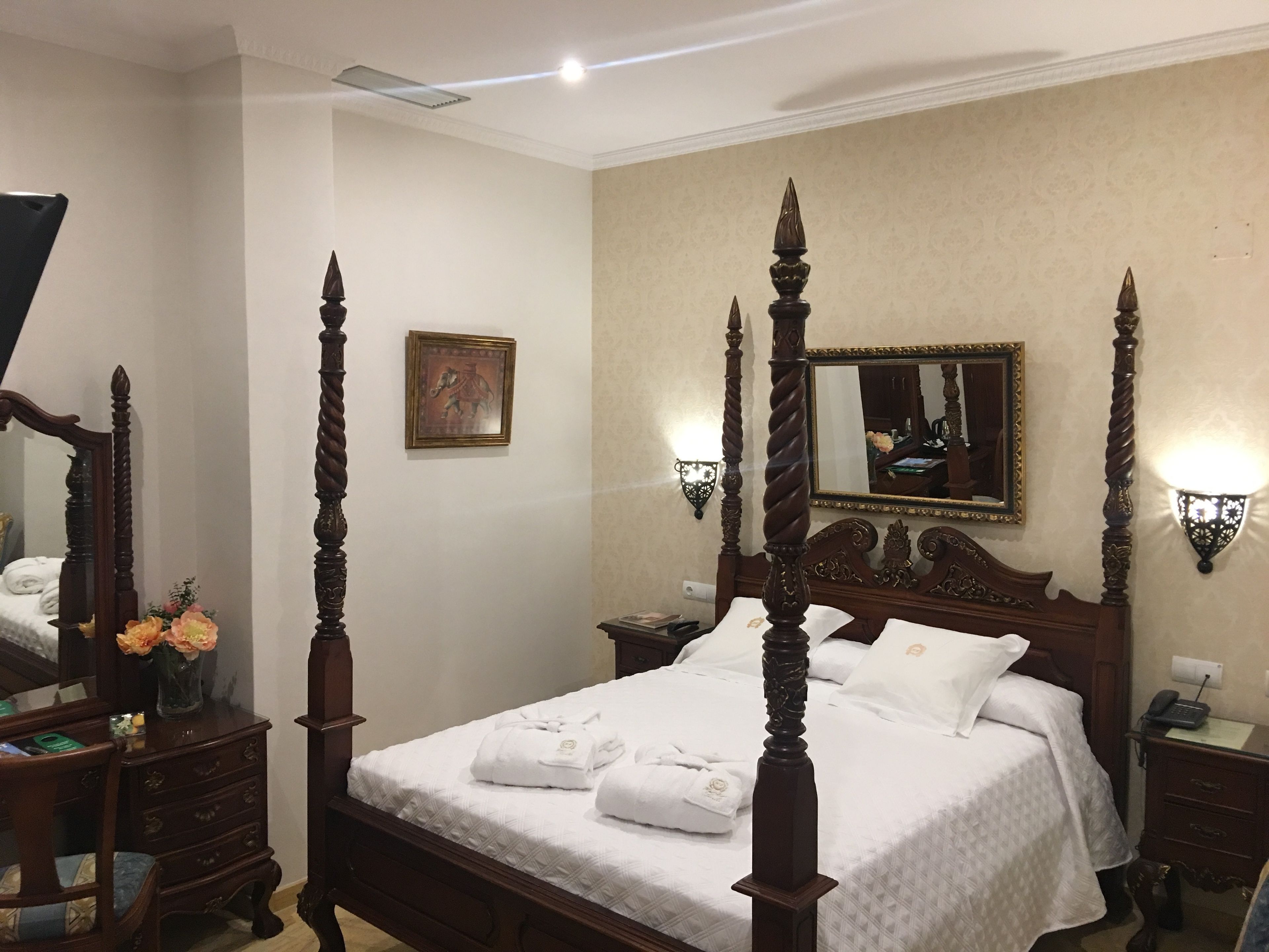 A four-poster bed with white linens, a bedside table with a lamp, and a mirror on the wall.
