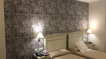 A hotel room with a patterned wallpaper depicting a cityscape, two beds with beige bedding, bedside tables with lamps, and a small desk with a phone and book.