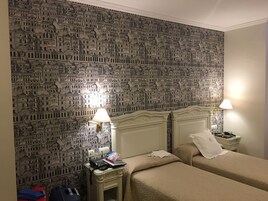 Una camera d'albergo con carta da parati decorata con un motivo cittadino, due letti con biancheria beige, comodini con lampade e una piccola scrivania con telefono e libro.