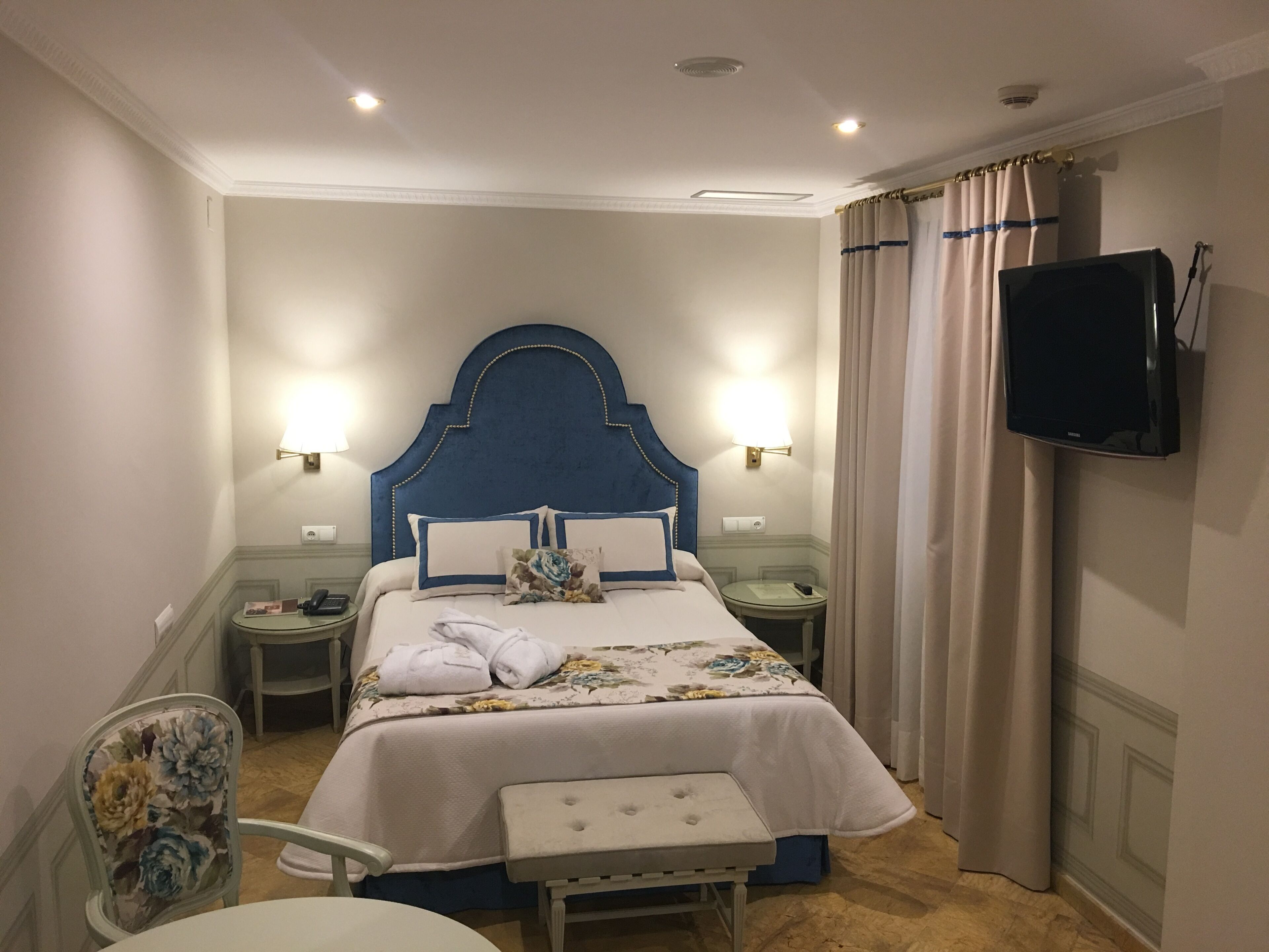 A hotel room with a bed, bedside lamps, a television, a chair, and a small table.