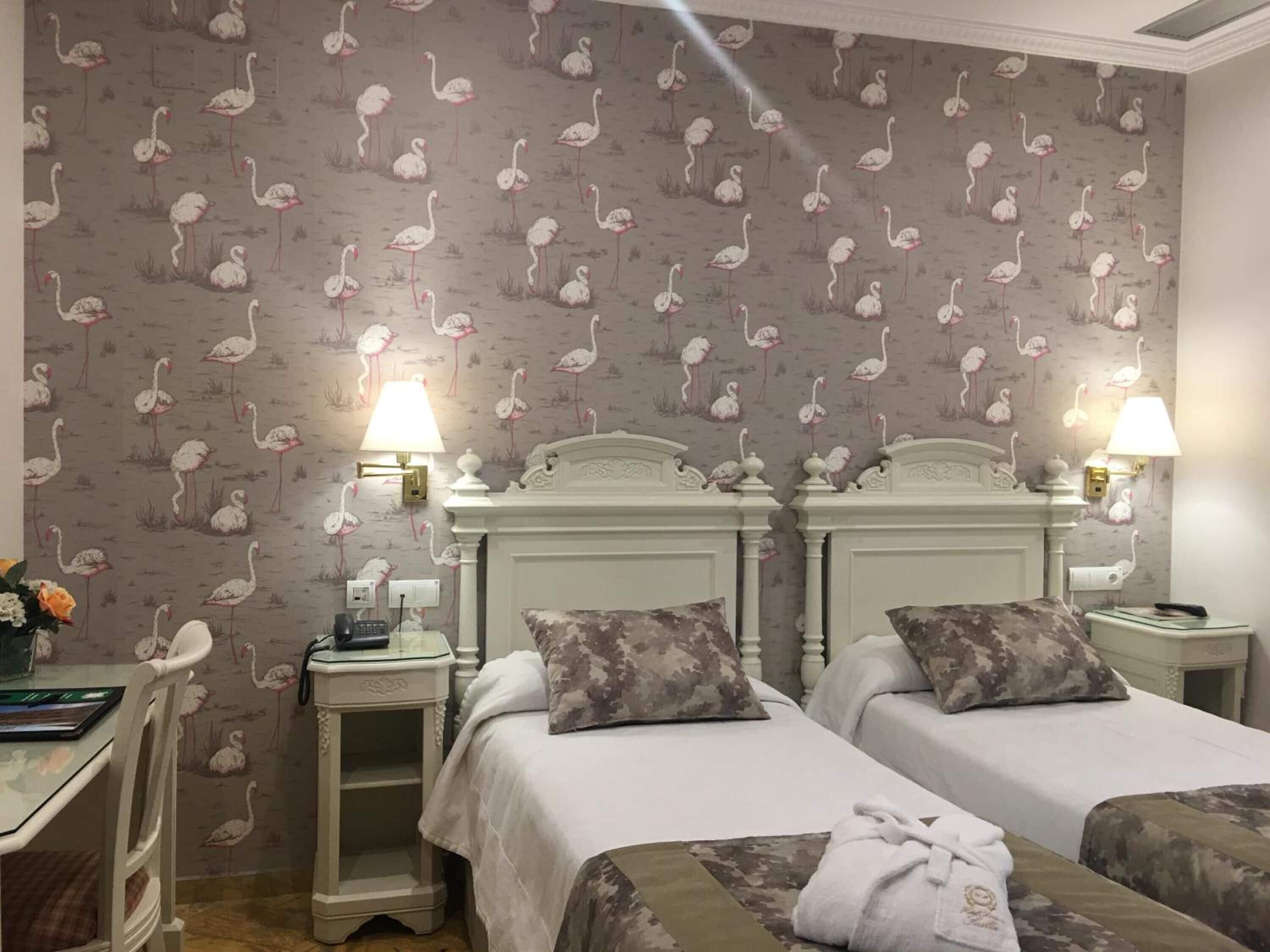 A hotel room with two beds, a nightstand with a lamp, a desk with a phone, and a wall with a flamingo pattern.