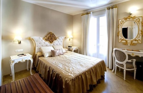 Une chambre d’hôtel classique, dotée d’un grand lit, de deux tables de chevet, d’un bureau avec une chaise, d’un miroir et d’une fenêtre avec des rideaux.