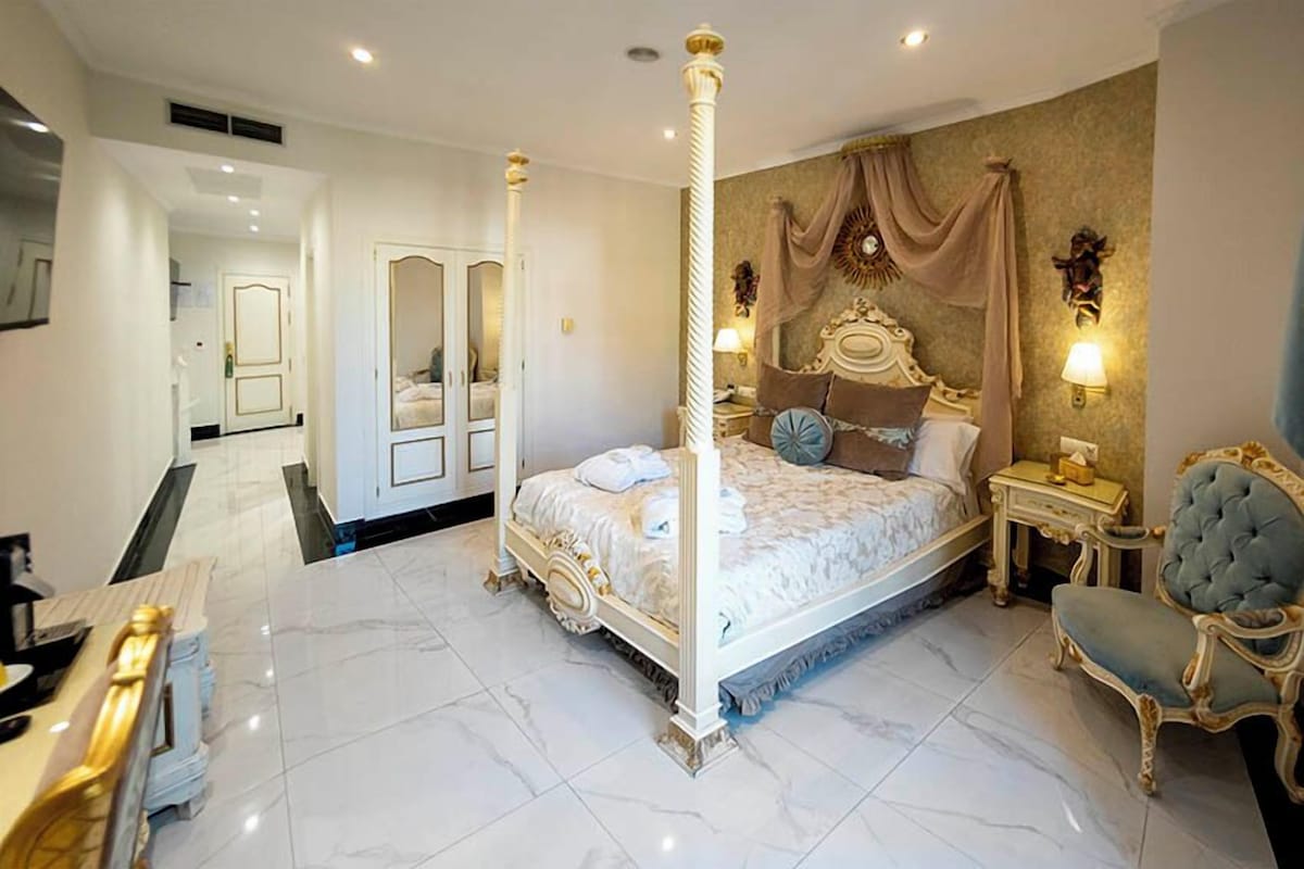 A four-poster bed with a canopy, a sitting chair, and a bedside table with a lamp.