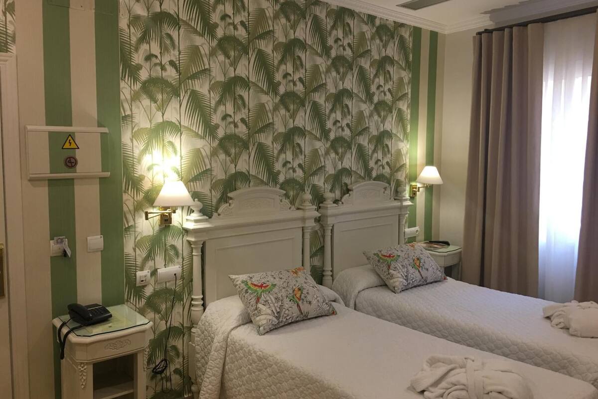 Une chambre d’hôtel avec deux lits, un mur à rayures vertes, un papier peint à motifs tropicaux et une fenêtre avec des rideaux.