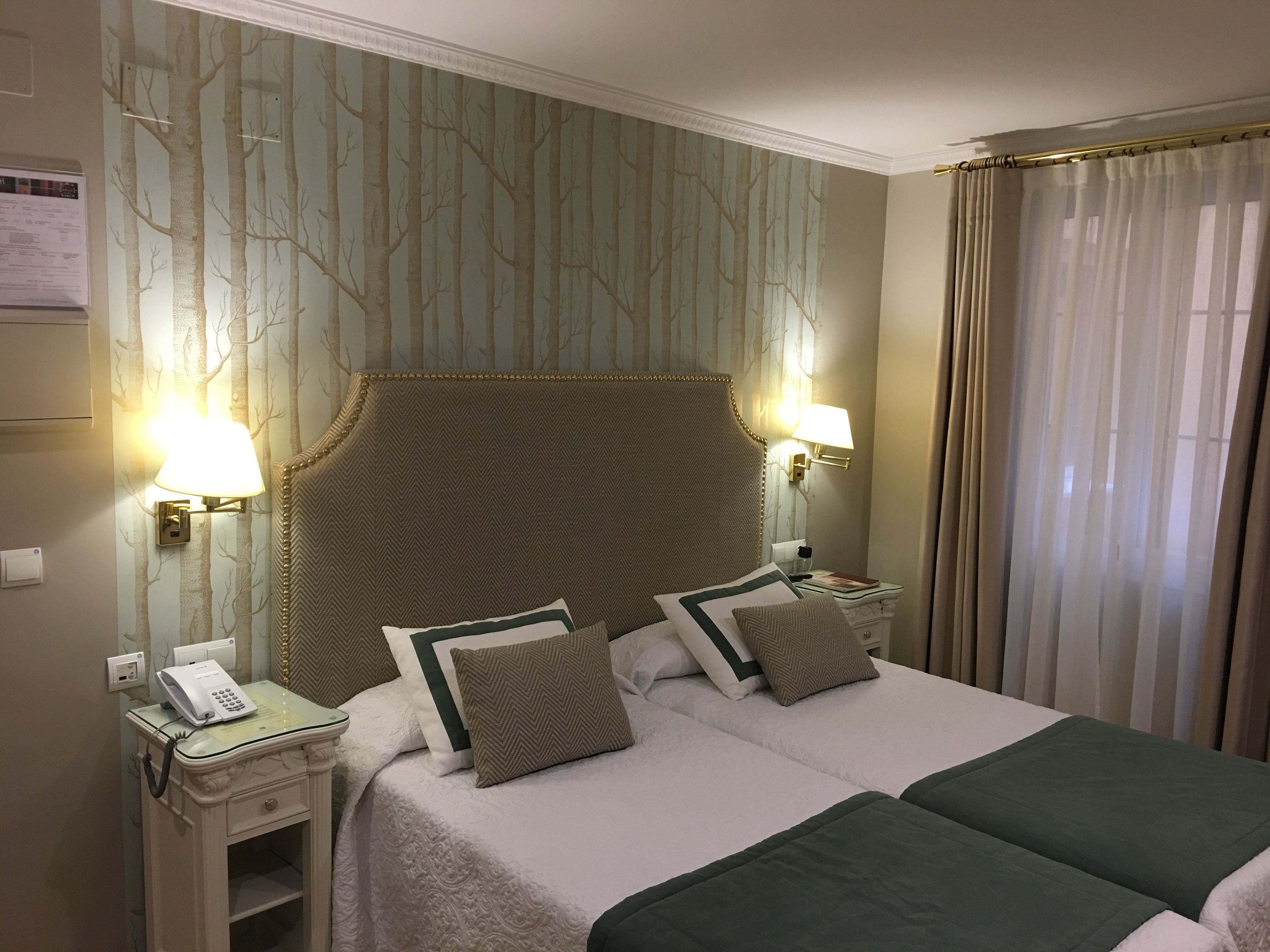 Habitación de hotel con dos camas, un cabecero grande, lámparas fijadas a la pared, una mesita de noche con teléfono y una ventana con cortinas.