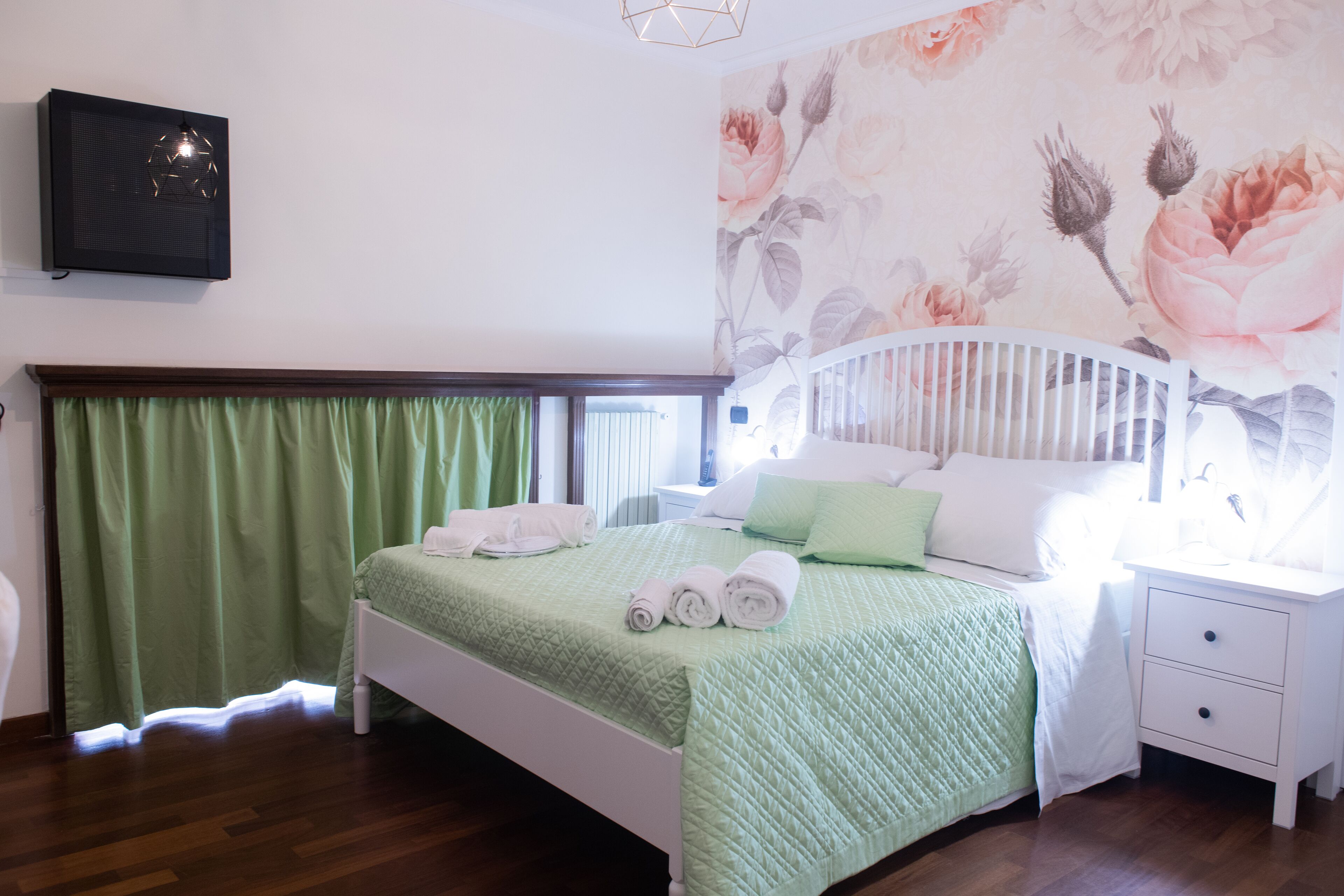Ein Schlafzimmer mit einem Bett, grünen Vorhängen, einem weißen Nachttisch und einer Wandmalerei mit Blumenmuster.
