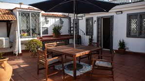 Une terrasse avec une table et des chaises en bois, entourée de plantes en pot et d’un grand parasol.