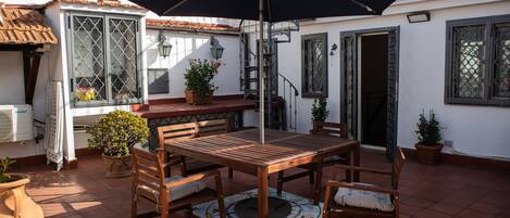 A patio with a wooden table and chairs, surrounded by potted plants and a large umbrella.