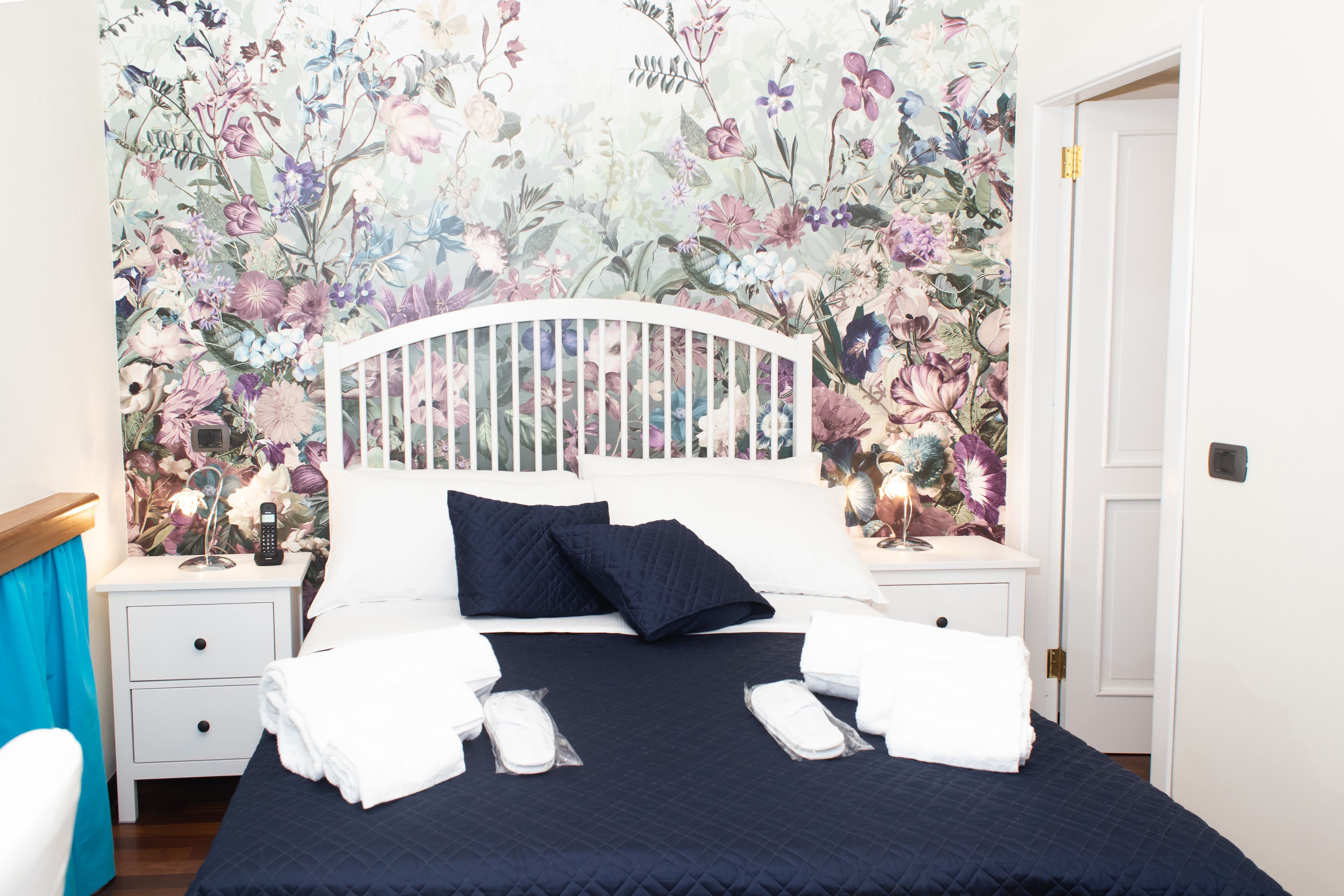 A neatly made bed with white linens and a dark blue quilt, flanked by two bedside tables with lamps, in a room with floral wallpaper.