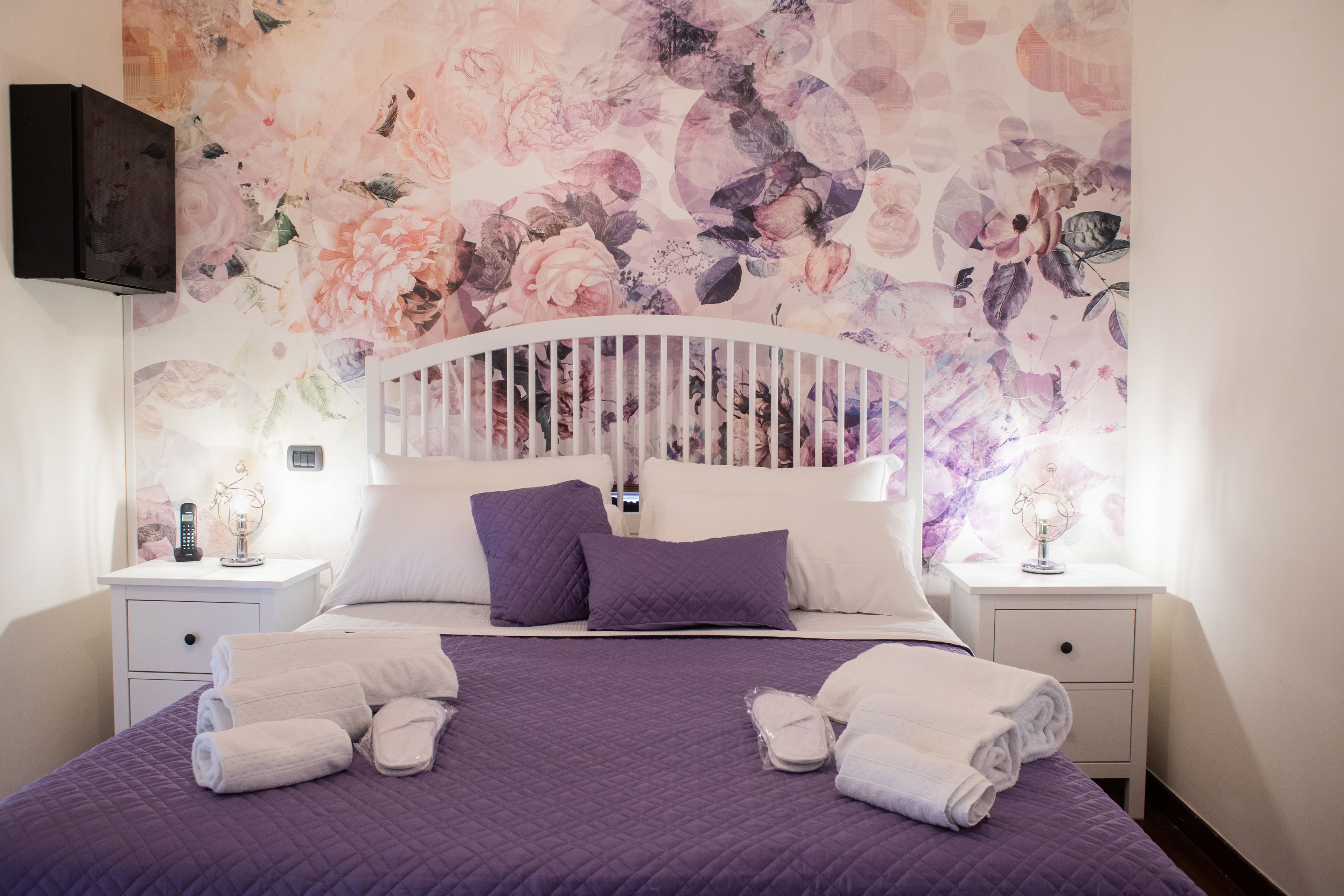 Ein ordentlich bezogenes Bett mit einer violetten Steudecke, weißen Kissen und gefalteten Handtüchern auf einem weißen Nachttisch.