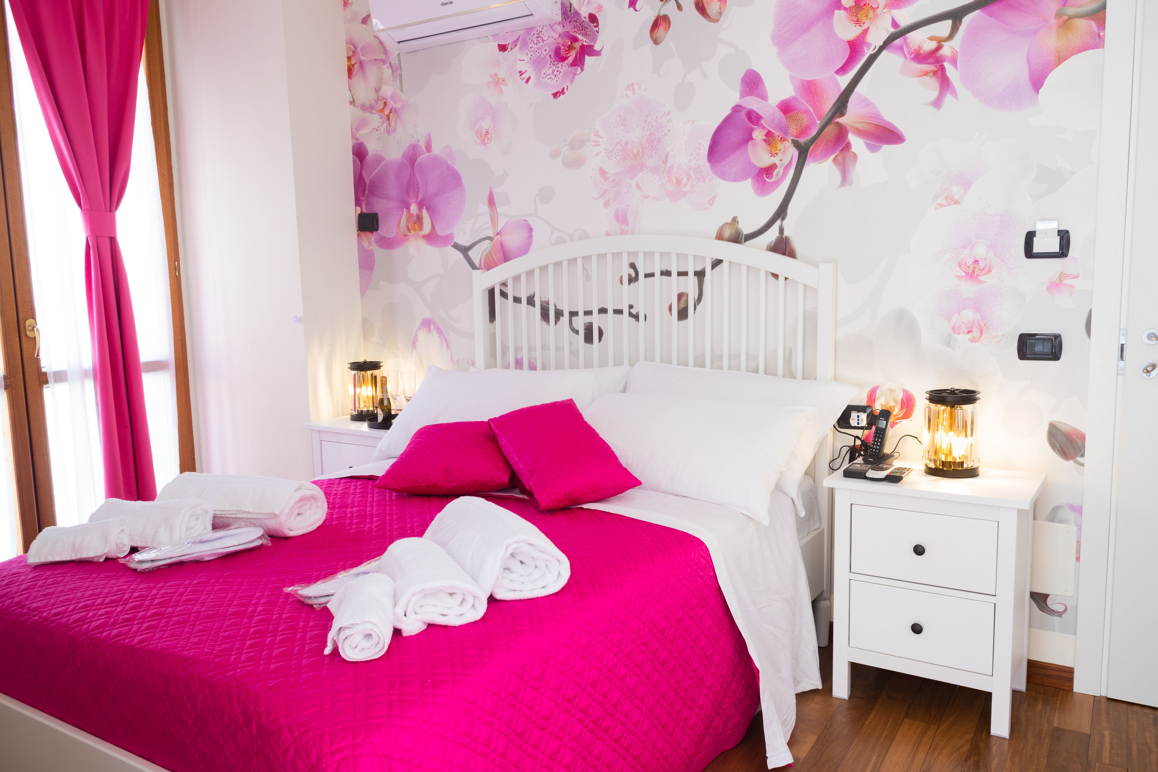 A hotel room with a bed, pillows, a nightstand, and a wall with floral wallpaper.