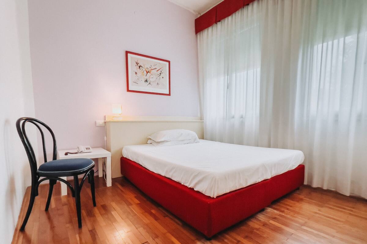 A hotel room with a wooden floor, a bed with a red base, a white headboard, a chair, a bedside table with a lamp, and a window with white curtains.