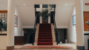 Una imponente escalera con alfombra roja, flanqueada por columnas, que conduce a una plataforma con una planta en maceta.