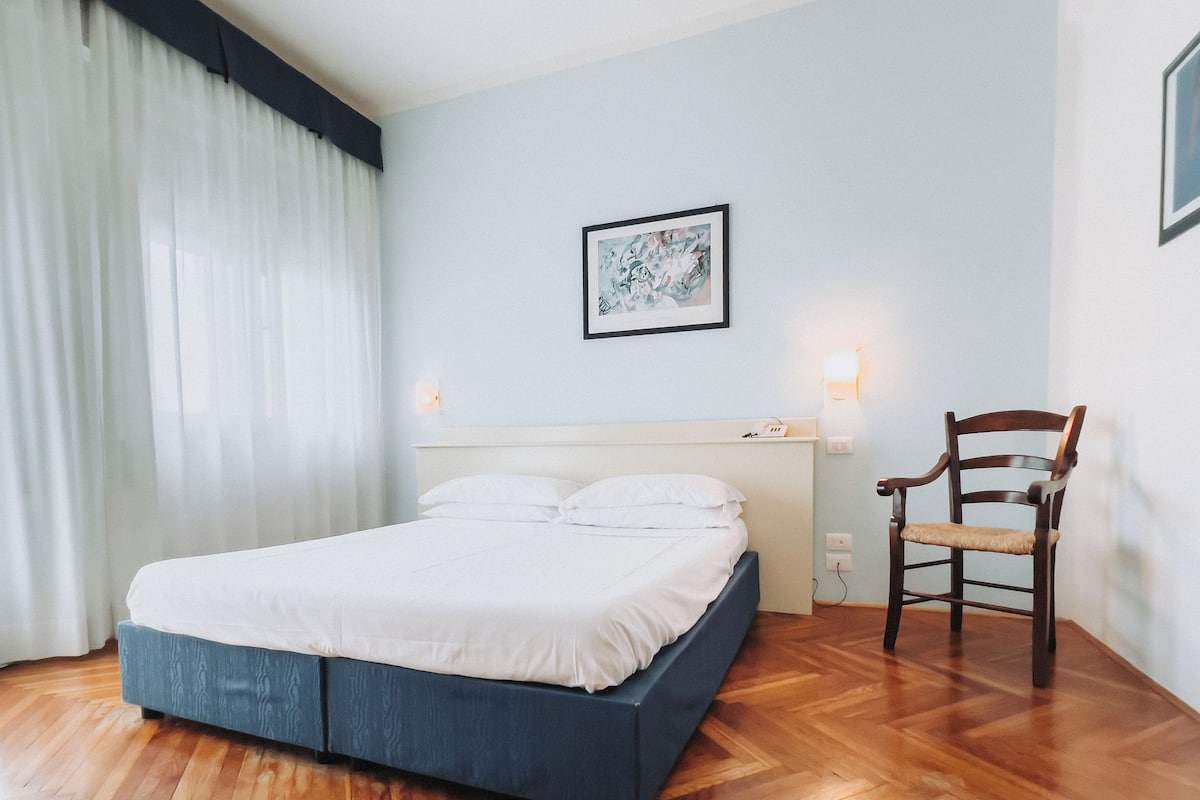 A neatly made bed with white linens in a room with wooden flooring, a wooden chair, and a framed artwork on the wall.