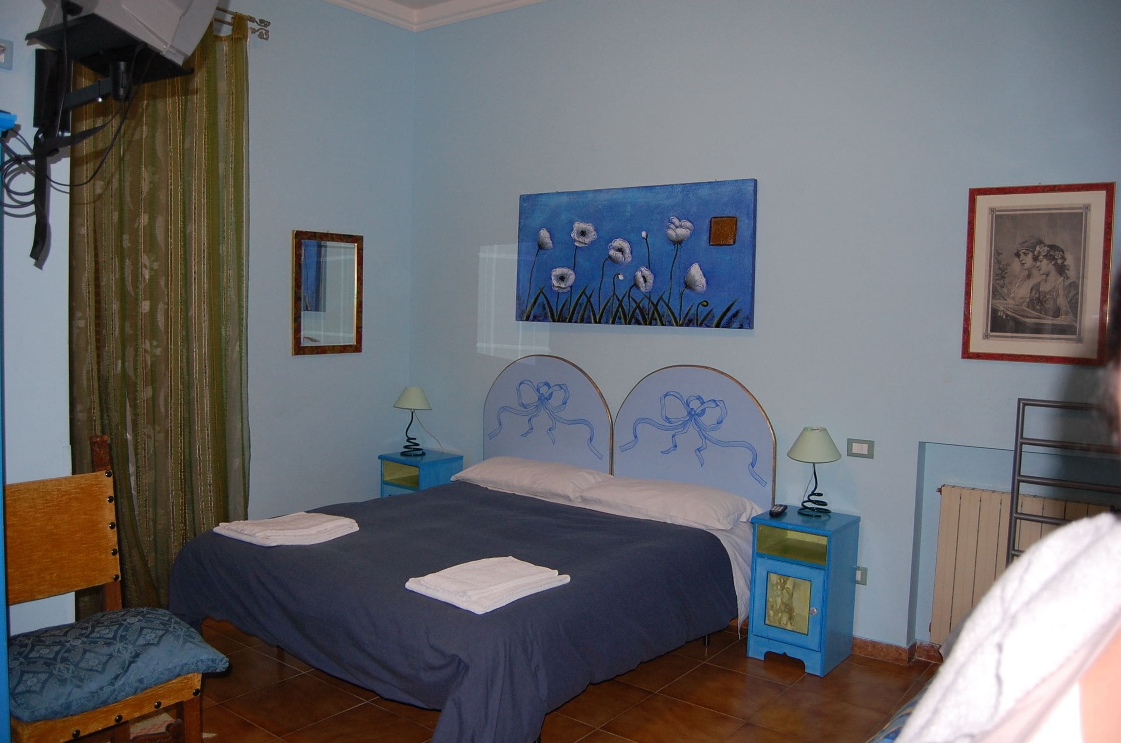 A bedroom with a bed, two bedside tables, a chair, a painting, and a framed picture on the wall.