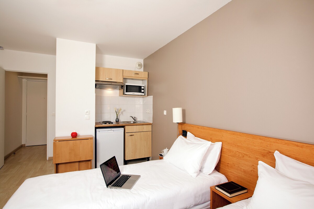 Une chambre d’hôtel avec un lit, une table de chevet sur laquelle se trouve un livre, un ordinateur portable, un four à micro-ondes dans la cuisine et une petite pomme rouge sur la table.