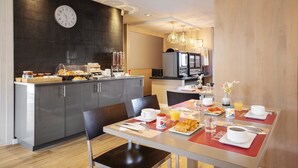 Een moderne hotel eetruimte met een keuken op de achtergrond, ontbijtartikelen op tafel en een klok aan de muur.