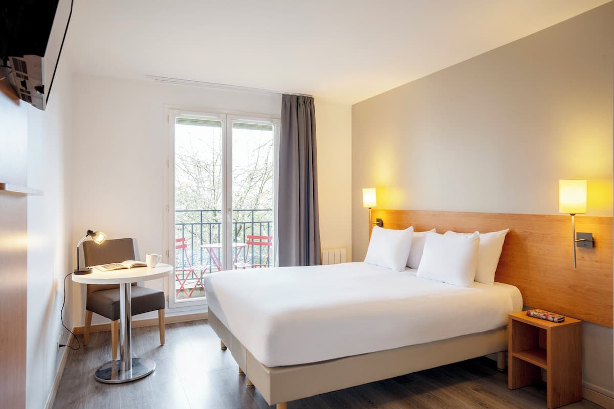 Ein Hotelzimmer mit einem großen Bett, Nachttischlampen, einem kleinen Tisch mit Stuhl und Blick auf den Balkon.